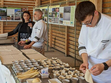 The image shows a culinary event where chefs and participants are preparing dishes made with white asparagus, with a large table full of ingredients and utensils.  A young chef in the foreground, wearing a white chef's jacket, is carefully folding a piece of puff pastry to create a culinary preparation.  On the table, several trays with fresh asparagus, pre-cut dough, and small metal molds are arranged, suggesting that tartlets or stuffed pastries are being prepared.  In the background, a man and a woman are speaking into a microphone, probably explaining the preparation steps or sharing the history of white asparagus.